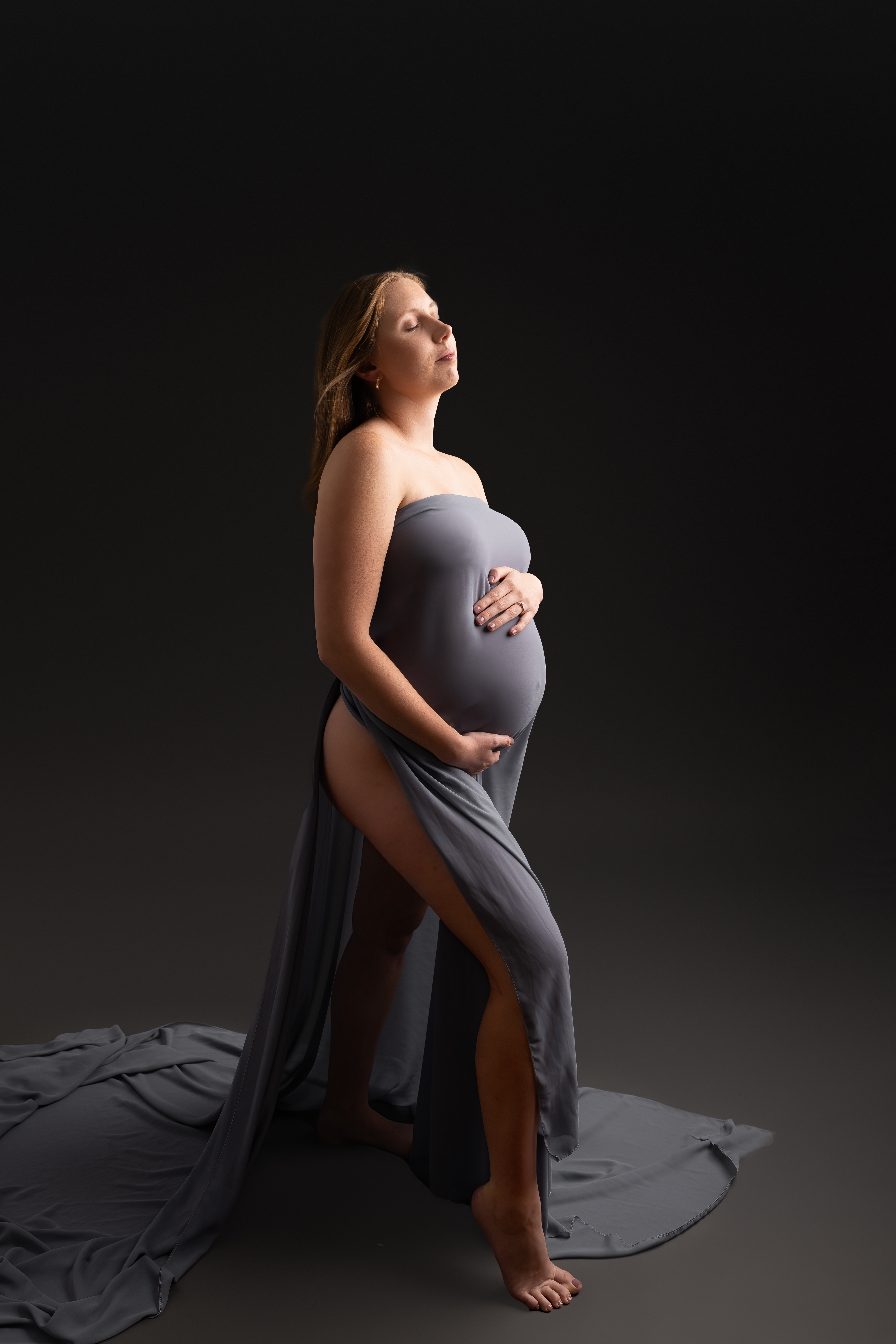 A pregnant woman in a grey dress against a dark background for Tampa maternity photography session