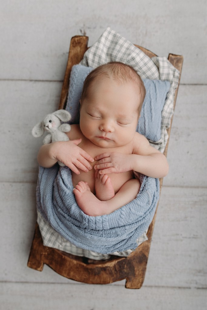 Newborn sleeping in blue wrap on rustic wooden prop against white backdrop.