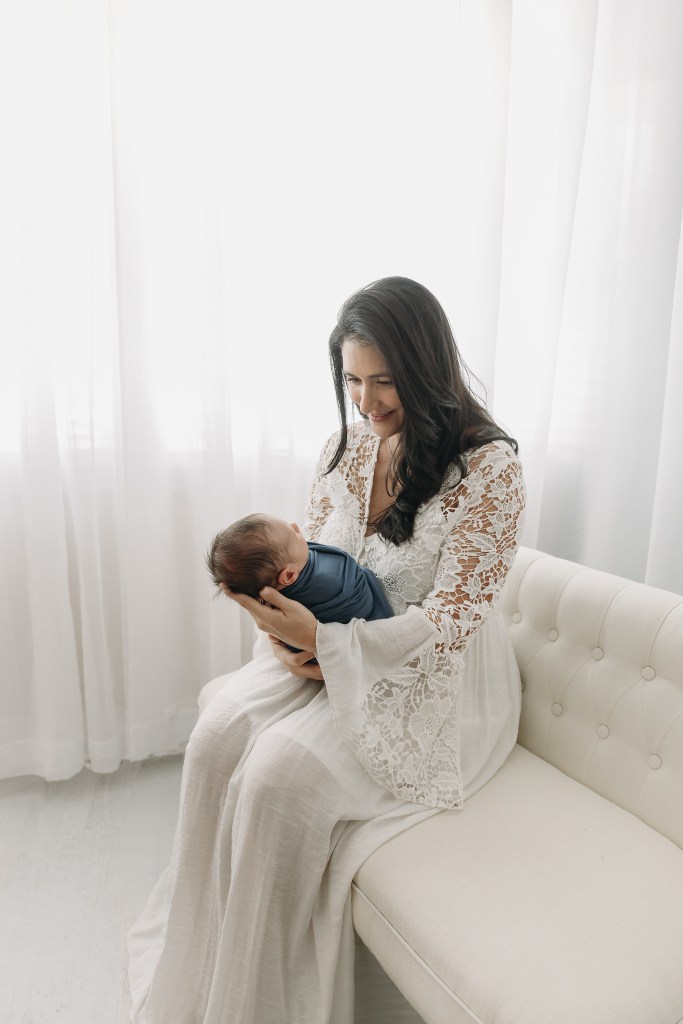 Mother in white lace dress sitting on a cream-colored couch holds her newborn baby 