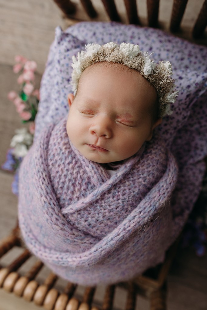 Sleeping newborn wrapped poses in soft purple knit blanket wearing delicate white floral crown