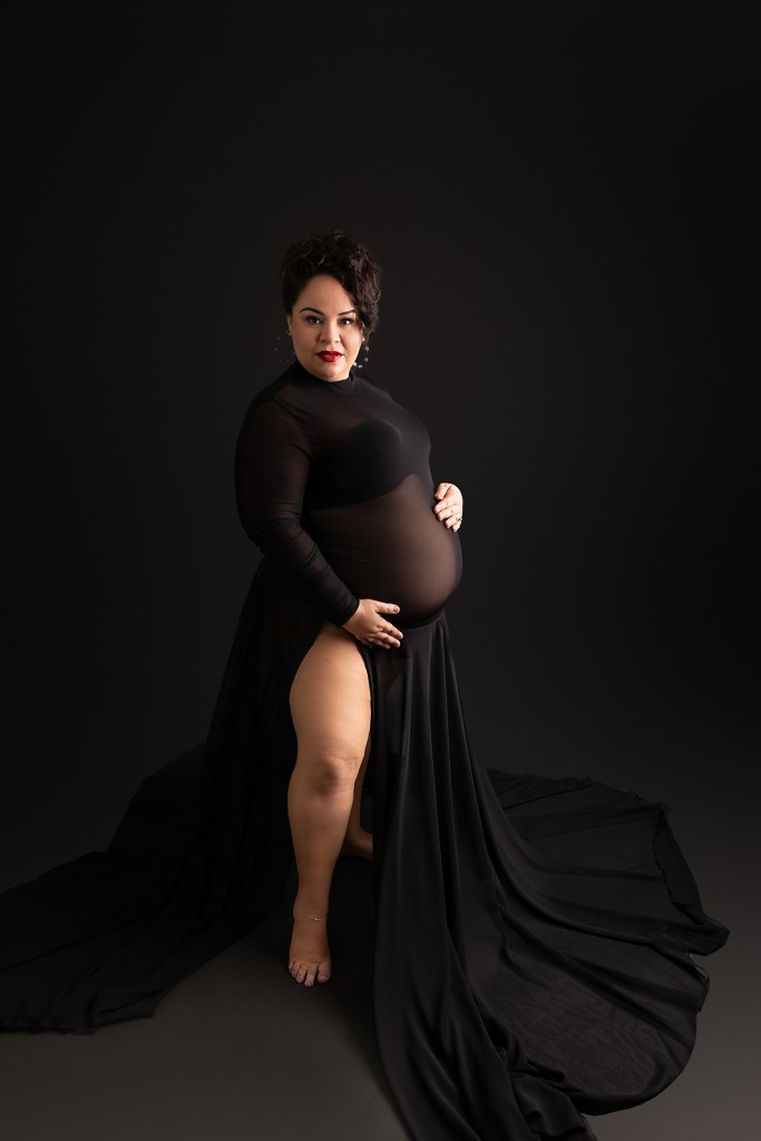 Artistic maternity portrait in black dress against dark background with flowing fabric and moody lighting 