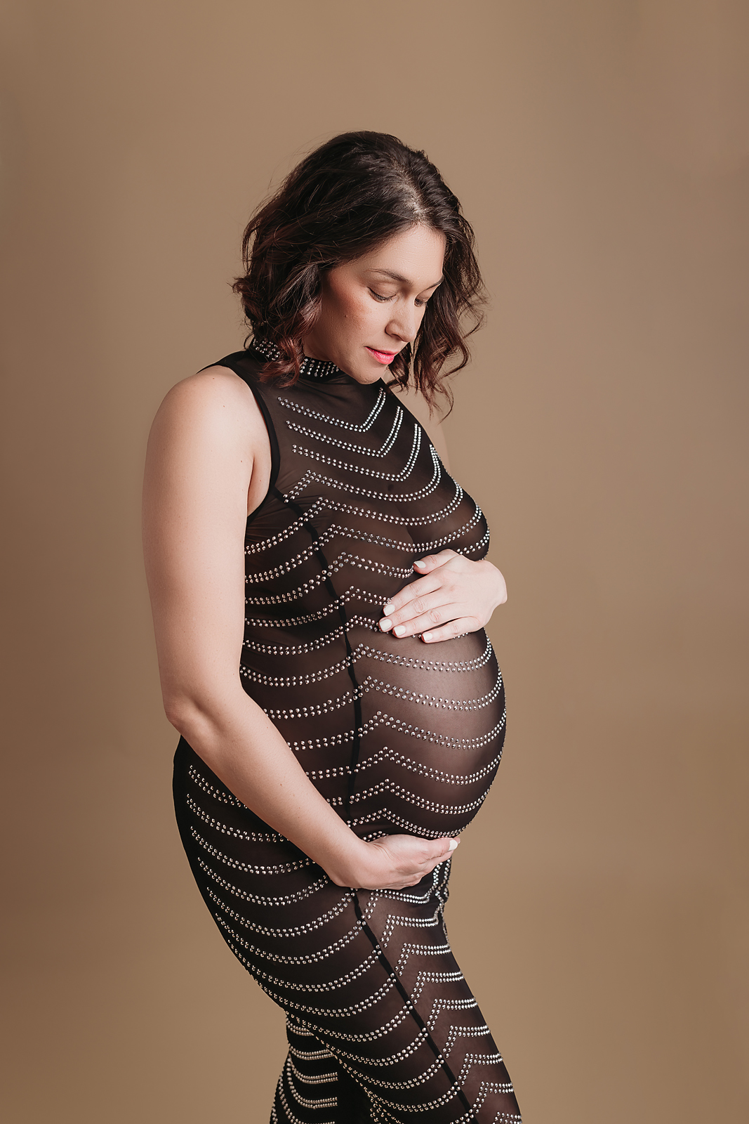 A pregnant woman in a striped mesh dress poses against a brown backdrop during a Tampa maternity photographer session