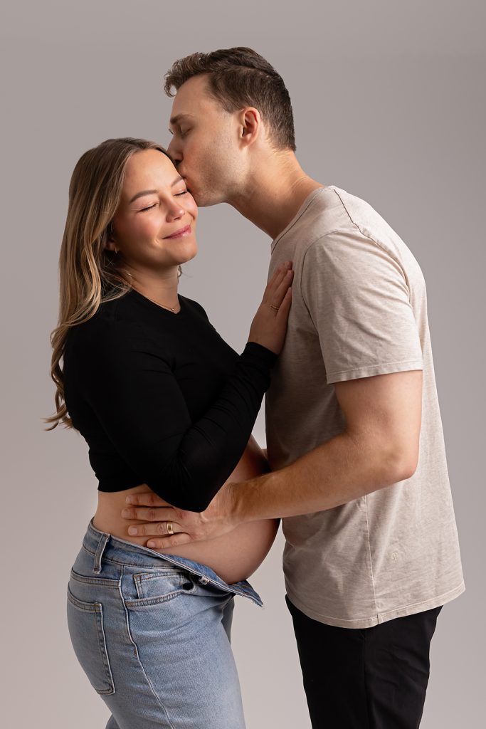 A pregnant couple embraces tenderly against a gray studio backdrop, wearing jeans and white top for maternity photos in Tampa