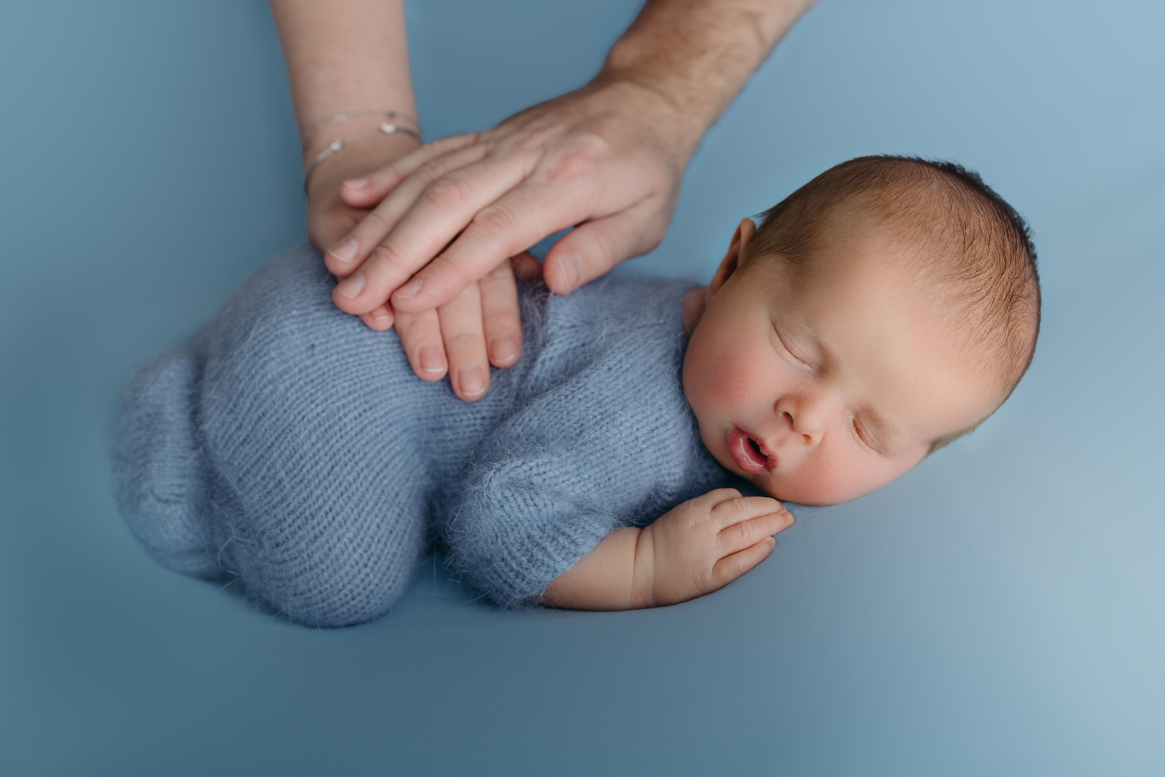 newborn baby boy in knit romper sleeps and mom and dad pat his back during tampa photographer session