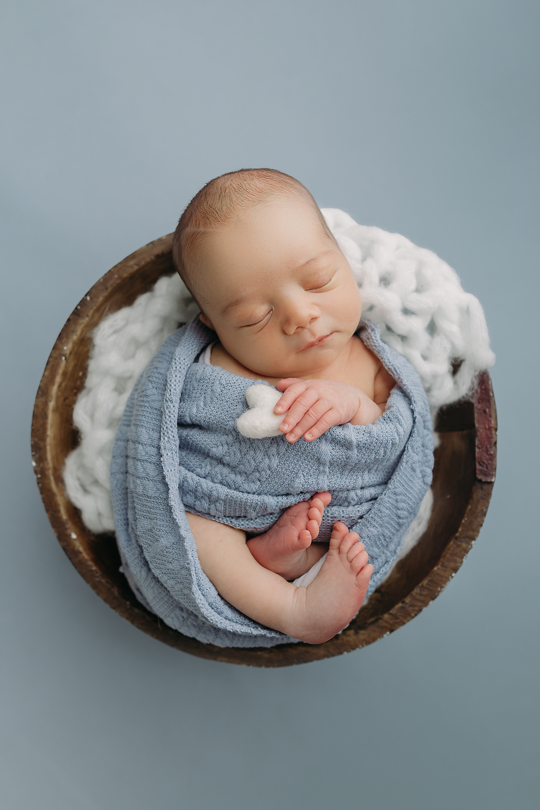 Peaceful sleeping newborn wrapped in soft blue swaddle rests in wooden bowl with white knit blanket for newborn photos in Tampa