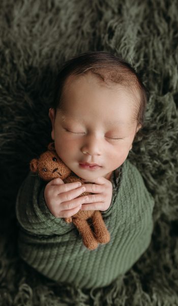 newborn baby boy swaddled in dark green holds teddy bear