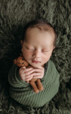 newborn baby boy swaddled in dark green holds teddy bear