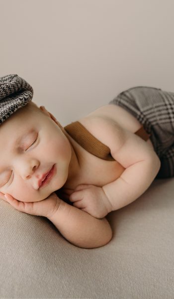 baby boy in overalls and hat poses for Tampa newborn photographer